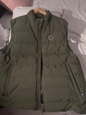 Olivgrön dunväst från Peak Performanc - Snygg olivgrön dunväst från Pelle p med hög krage och dragkedja framtill. Västen har två snedställda fickor med dragkedja och en diskret logotyp på bröstet. Perfekt för lager-på-lager och streetwear.