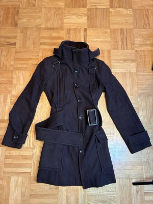 Mörkbrun trenchcoat med bälte - Snygg mörkbrun trenchcoat med hög krage och brett bälte i midjan. Jackan har stora knappar framtill, dekorativa axelband och fickor med lock. Tillverkad i ett kraftigt tyg som ger en stilren och modern look.