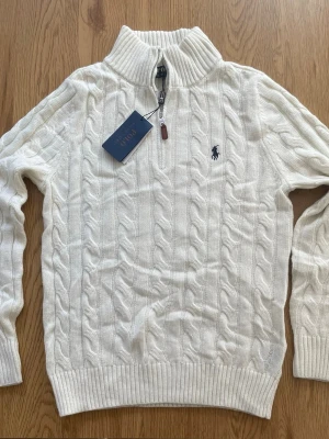 Vit kabelstickad tröja Polo Ralph Lauren - Vit kabelstickad tröja från Polo Ralph Lauren med halv dragkedja och hög krage. Tröjan har ribbstickade muddar och nederkant samt den klassiska broderade loggan på bröstet. Perfekt för lager-på-lager och snygg till jeans.