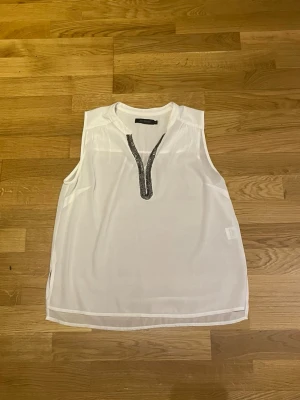 Vit blus från Vera Moda med pärldetaljer - Stilren vit blus i lätt och luftig polyester med ärmlös design. Blusen har en v-ringad öppning framtill med dekorativa detaljer längs kanten som ger en extra touch. Perfekt att matcha med jeans eller kjol.