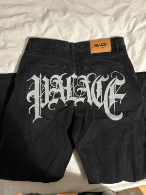 Svarta Palace jeans med stor logga - Säljer ett par svarta jeans från Palace med stor vit logga i gotisk stil tryckt över bakfickorna. Klassisk femficksmodell med läderpatch i midjan där det står 'Palace London'. Perfekta för dig som gillar streetwear och vill sticka ut.