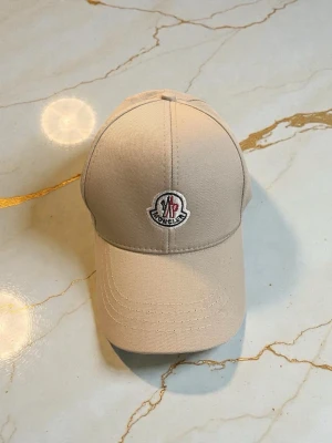 Beige keps från Moncler - Snygg beige keps från Moncler med klassisk böjd skärm och broderad Moncler-logga framtill. Tillverkad i slitstark bomull med justerbar rem bak för perfekt passform. Enkel och stilren design som passar till det mesta.