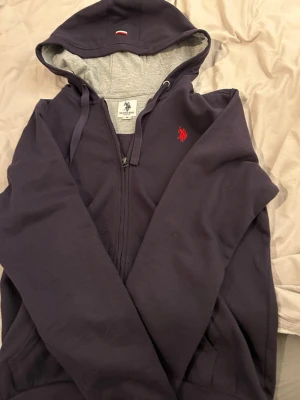 Mörkblå hoodie från Polo Assn  - Mörkblå hoodie från Polo Assn med dragkedja och snörning i huvan. Klassisk röd broderad logga på bröstet och mjukt grått innerfoder i huvan. Perfekt för en avslappnad och stilren look.