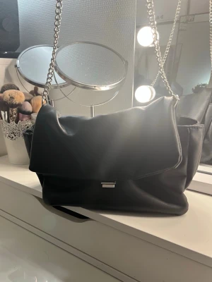 Svart axelväska med kedjerem - Stilren svart axelväska i mjukt konstläder med bred lockflik och silverfärgad kedjerem. Väskan har en enkel metallknäppe framtill och rymlig insida. Perfekt för dig som gillar en clean och modern look.