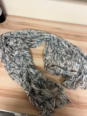 Mönstrad halsduk från Name It - Ljus halsduk från Name It med ett snyggt blad- och grenmönster i grått, svart och blått. Tillverkad i ett tunt och mjukt material som känns skönt mot huden. Perfekt accessoar för att lyfta din outfit med lite extra stil.