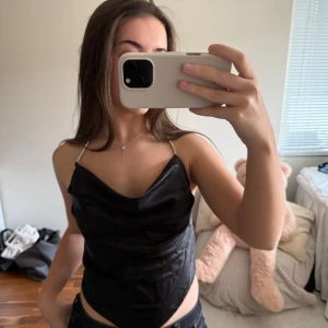 Svart satin linnetopp med smala band - Snygg svart linnetopp i satin med smala axelband och draperad urringning. Toppen har en croppad passform och ger en elegant look. Perfekt att styla med jeans eller kjol för en trendig outfit.