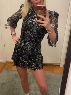 Svart och silver glittrig kortklänning - Snygg svart kortklänning med silvrigt glittrigt mönster och omlottknytning i midjan. Klänningen har trekvartsärmar i transparent spets och volangdetaljer vid ärmslut. Perfekt för dig som vill sticka ut med en trendig look.