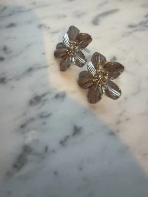 Blommiga örhängen i silverfärg - Säljer ett par snygga örhängen i form av blommor med detaljerade blad. De är i silverfärg och har en stilren, modern look som verkligen sticker ut. Perfekta för dig som vill ha något unikt och trendigt till din outfit. Aldrig använda