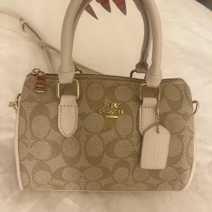 Coach beige mini rowan crossbody bag - Snygg beige miniväska från Coach med klassiskt monogrammönster och guldfärgade detaljer. Väskan har två korta handtag, avtagbar axelrem och dragkedja upptill. 