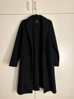 Svart kappa från Zara med klassisk krage - Stilren svart kappa från Zara med rak passform och klassisk krage. Kappan har långa ärmar och är tillverkad i ett mjukt tyg som ger en clean look. Perfekt för dig som gillar minimalistisk stil och vill ha en enkel men snygg kappa till garderoben.