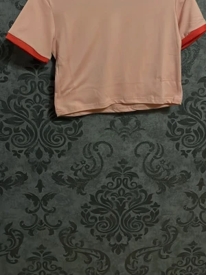 Rosa cropped t-shirt med röd kant - Snygg rosa cropped t-shirt med kontrasterande röd kant vid ärmslut och halsringning. T-shirten har en enkel och modern design, perfekt för en avslappnad stil. Tillverkad i mjukt material som känns skönt mot huden.