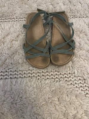 Gröna sandaler med remmar - Snygga sandaler med gröna textilremmar i korsad design och beige sula. Remmarna har små vita prickar och sandalerna har öppen tå samt justerbar knytning vid ankeln. Perfekta för sommaren och ger en avslappnad vibe. Använda men väldigt bekväma 
