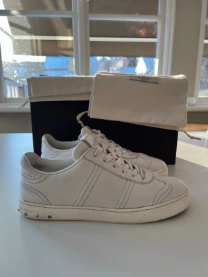 Vita Valentino Garavani Flycrew - Vita valentino Garavani Flycrew-sneakers i slätt läder med låg profil och stilren design. Vita snören, diskreta sömmar och nitar på hälen, en nit har lossnat men medföljer och kan enkelt limmas tillbaka. Skorna är i mycket bra skick. Storlek 39,5 men de är stora i storleken så passar ca 40-41. 100% äkta, köpta via Vestiare Collective. Perfekta för dig som gillar stilrena och tidlösa plagg. Obs att endast skor medföljer! Vid frågor eller funderingar är det bara att höra av sig. Mvh Olof.