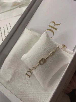 Guldfärgat armband från Dior med pärlor - Snyggt armband från Dior i guldfärgad metall med små vita pärlor och gnistrande stenar. Armbandet har en tunn kedja och ett dekorativt CD-emblem i mitten. Perfekt accessoar för att lyfta din stil.