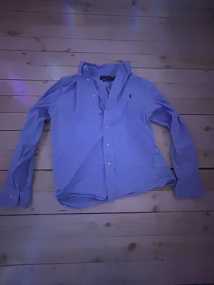 Blå skjorta från Ralph Lauren - Klassisk blå skjorta från Ralph Lauren med lång ärm och knappar framtill. Skjortan har den ikoniska broderade loggan på bröstet och är tillverkad i bomull. Perfekt för en clean och stilren look.säljer denna skjortan för att den blivit för liten.nypris cirka 1000kr