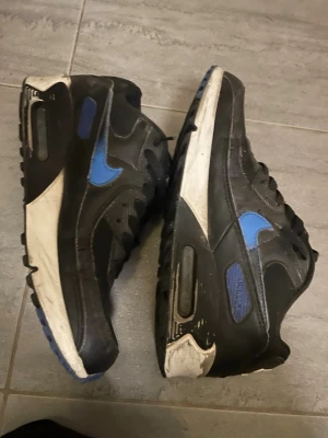 Nike Air Max 90 svart/blå sneakers - Säljer ett par Nike Air Max 90 sneakers i svart med blå detaljer och vit sula. Klassisk modell med synlig Air-enhet i hälen, ovandel i läder och mesh samt ikonisk Swoosh på sidan. Perfekta för dig som gillar streetwear och sportig stil.