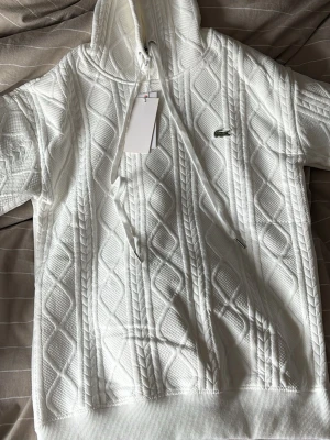 Vit hoodie från Lacoste  - Säljer en vit hoodie från Lacoste med snyggt strukturmönster i kabelstickad stil. Hoodien har dragsko i huvan, ribbade muddar och den klassiska gröna krokodilloggan på bröstet. Perfekt för dig som gillar stilrena och sportiga plagg.