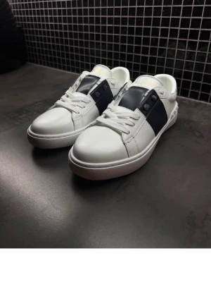 Valentino opens - Snygga vita sneakers med mörkblå panel på sidan och klassisk snörning. Skorna har en stilren design och är tillverkade i syntetmaterial med vit sula och diskreta detaljer. Perfekta för dig som gillar en clean look.
