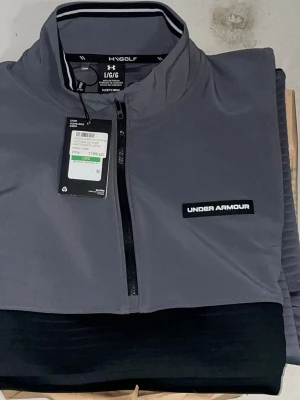 Grå half-zip tröja från Under Armour - Snygg grå tröja från Under Armour med svart panel över bröstet och halv dragkedja. Tröjan har hög krage, lång ärm och logga på bröstet. Perfekt för dig som gillar sportig och clean stil.