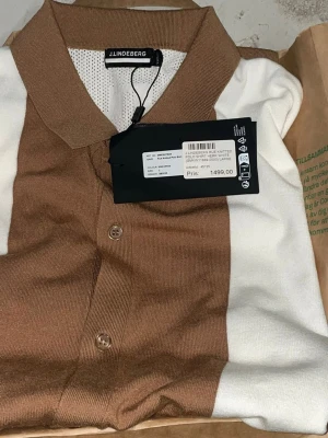 Brun och vit skjorta från J.Lindeberg - Kortärmad skjorta från J.Lindeberg i brunt och vitt med knappar framtill. Skjortan har klassisk krage och är tillverkad i ett stickat material som ger en modern och stilren look. Perfekt för dig som gillar en clean och trendig stil.