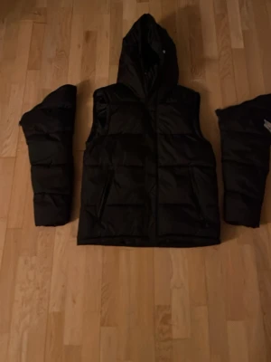 Burberry Svart dunjacka  - Snygg svart dunjacka med huva och avtagbara ärmar, vilket gör att den kan bäras både som jacka och väst. Jackan har quiltad design och dragkedja framtill. Perfekt för kalla dagar och flexibel stil.