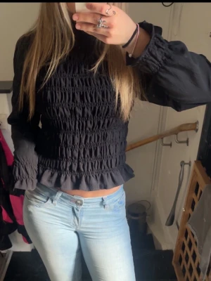 Svart smockad blus med volang - Trendig svart blus med smockad kropp och volangkant nedtill. Långärmad modell med lätt puff vid axlarna och resår vid ärmslut. Storlek S men passar upp till L då den är väldigt stretchig!