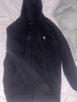 Svart hoodie från Polo Ralph Lauren M/S - Säljer en svart hoodie med dragkedja från Polo Ralph Lauren. Bra skick inte trasig bara lite tappad färg. Klassisk design med vit broderad logga på bröstet, huva med snörning och två fickor framtill. 