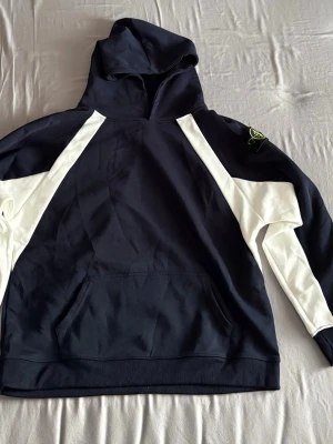 Stone Island marinblå hoodie - Snygg marinblå hoodie från Stone Island med vita kontrastpaneler längs ärmarna och axlarna. Klassisk huva och känguruficka framtill. Ikonisk Stone Island patch på vänster ärm. Tillverkad i mjuk bomull för skön känsla. Fick den som present men den var tyvärr för stor. Priset kan sänkas vid snabbaffär. Vid frågor är det bara att skicka ett meddelande så svarar jag så fort som möjligt.