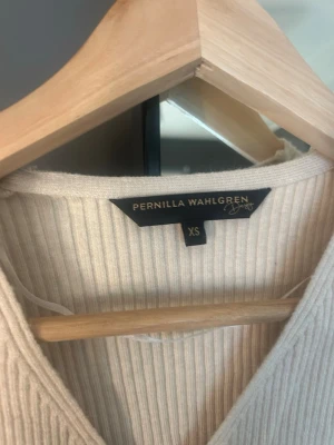 Ribbad omlottopp från Pernilla Wahlgren - Snygg ribbad omlottopp i krämvit färg från Pernilla Wahlgren. Tröjan har lång ärm, V-ringning och figurnära passform. Aldrig använd