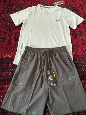 Under Armour träningsset t-shirt & shorts - Snyggt träningsset från Under Armour med en ljusgrå t-shirt och mörkgrå shorts. T-shirten har rund hals och logga på bröstet, medan shortsen har resårmidja, dragsko och sidofickor. Perfekt för gymmet eller löpning.