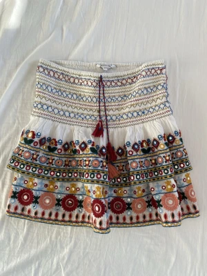Vit broderad kortkjol från Ba&sh - Supersöt vit kortkjol från zara med färgglada broderier i rött, blått, gult och grönt. Kjolen har volang, snörning med tofsar och ett unikt mönster med blommiga detaljer. Som man ser på sista bilden har sömarna gått upp lite, annars bra skick😁