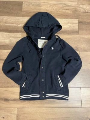 Abercrombie & Fitch hoodiejacka - sitter som M - Clean mörkblå hoodiejacka/cardigan från Abercrombie & Fitch. Muscle fit som sitter riktigt snyggt – upplevs som Medium. Bra skick, inga defekter. Tidlös design med klassisk A&F-logga.  📏 Mått: • Längd: 68 cm • Bredd/midja: 44 cm  Perfekt för vardag, skola & chill. Hör av er vid frågor!