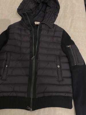 Svart dunjacka med huva från Moncler - Snygg svart dunjacka från Moncler med huva, ribbade muddar och ärmar i stickat material. Jackan har två framfickor med tryckknappar och en extra ficka med dragkedja på ärmen. Perfekt för kalla vinterdagar och riktigt trendig look.