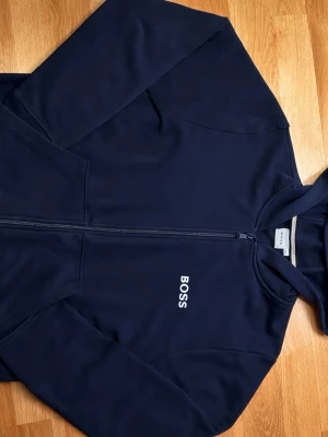 BOSS hoodie – mörkblå -  Snygg och stilren hoodie från BOSS i mörkblå färg. Klassisk design med diskret BOSS-logga på bröstet och hel dragkedja. Skön passform och mjukt, högkvalitativt material som funkar perfekt både till vardag och chill.  ✔️ Mycket bra skick ✔️ Inga fläckar eller hål ✔️ Från rök- och djurfritt hem ✔️ Äkta BOSS  Perfekt hoodie för dig som vill ha något stilrent men ändå bekvämt. Skicka gärna meddelande vid frågor eller om du vill ha fler bilder!