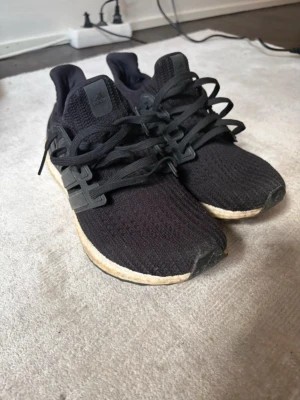Svarta Adidas Ultraboost sneakers - Svarta Adidas Ultraboost sneakers med stickad ovandel och klassiska tre ränder i plast på sidorna. Skorna har en vit Boost-sula som ger grym komfort och svarta skosnören. Perfekta för dig som gillar sportig och stilren look.