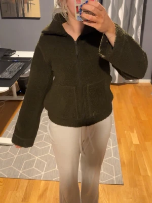 Mörkgrön teddyjacka med dragkedja - Mysig mörkgrön teddyjacka med hög krage och hel dragkedja framtill. Jackan har två stora fickor fram och är gjord i fluffigt teddy-material som håller dig varm. Rak passform och långa ärmar ger en avslappnad look.