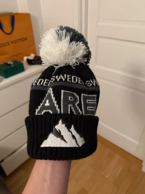 Svart och vit Åre mössa med tofs - Säljer en svart och vit mössa från Åre med stor tofs på toppen. Mössan har ett broderat bergmotiv framtill och texten 'SWEDEN' samt 'ÅRE' runt om. Ribbstickad kant och etikett bak. Perfekt för kalla dagar och snygg streetstil.
