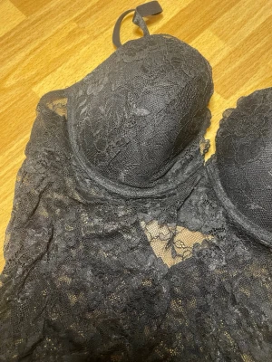 Svart spetsbody med vadderade kupor - Snygg svart body i genombruten spets med vadderade kupor och justerbara axelband. Bodyn har ett feminint blommönster och en liten cut-out detalj framtill. Perfekt för dig som vill ha något extra under kläderna. HELT NY!!! Utan lapp.