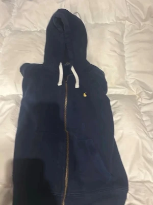 Mörkblå hoodie från Polo Ralph Lauren - Mörkblå hoodie med dragkedja från Polo Ralph Lauren. Klassisk design med vit dragsko i huvan och gul broderad logga på bröstet. Tillverkad i mjukt bomullsmaterial och har kängurufickor framtill. Perfekt för en avslappnad och stilren look.
