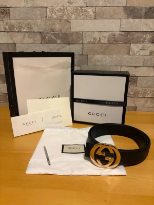Svart Gucci bälte med guldfärgat spänne - Säljer ett svart bälte från Gucci med det ikoniska GG-spännet i guld. Bältet är gjort i skinn och kommer med originalpåse, box och påse från Gucci. Perfekt accessoar för att lyfta vilken outfit som helst. Hålgörare medkommer ifall bältet inte sitter exakt så som man tänkt, allt efter kundens behov!