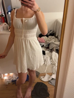 Vit kort sommarklänning med knyt - Söt vit kort klänning med knyt framtill och breda axelband. Klänningen har en lätt rynkad struktur och smock i midjan för en bekväm passform. Perfekt för varma dagar och enkel att styla med accessoarer.