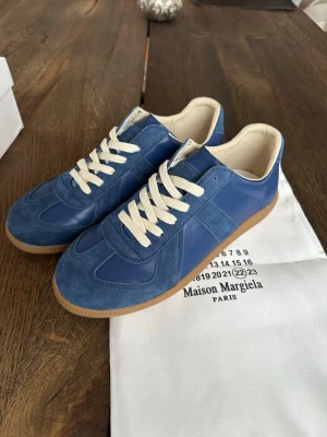 Blå Maison Margiela sneakers - Snygga blå sneakers från Maison Margiela med ovandel i mocka och läder, vita snören och klassisk gummisula. Skorna har en låg silhuett och stilren design som passar perfekt till streetwear och casual outfits.