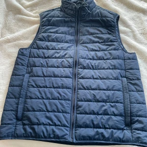 Mörkblå quiltad dunväst - Snygg mörkblå dunväst med quiltat mönster och hög krage. Västen har dragkedja framtill, två sidofickor och en innerficka. Perfekt för lager-på-lager och ger en clean look. Materialet är lätt och smidigt, vilket gör den enkel att bära till olika outfits.