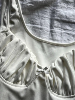 Vit kort nattlinne med rosett - Söt vit bodyconklänning med rynkade kupor och liten rosett framtill. Klänningen har breda axelband och är tillverkad i ett stretchigt, mjukt material som sitter tajt mot kroppen. Perfekt för en trendig och stilren look. Köpt på marknad i Spanien, vet ej märke. 