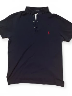 Marinblå pikétröja från Polo Ralph Lauren - Klassisk marinblå pikétröja från Polo Ralph Lauren med korta ärmar, krage och knappar framtill. Ikoniska röda logobroderiet på bröstet. Tillverkad i mjuk bomull som känns skön mot huden. Perfekt till jeans eller shorts för en clean look.