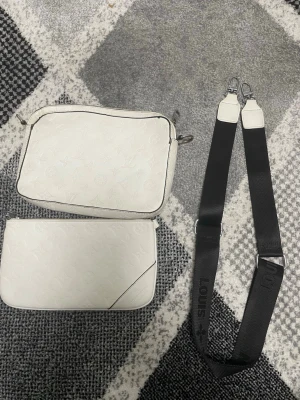 Vit Louis Vuitton handväska med axelrem - Säljer en vit handväska från Louis Vuitton med klassiskt monogrammönster i präglat läder. Väskan har en avtagbar svart axelrem med 'LOUIS' och monogramdetaljer. Två separata fack ingår, perfekt för att organisera dina grejer.