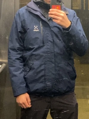 Blå vindjacka från Haglöfs - Snygg mörkblå vindjacka från Haglöfs med dragkedja och hög krage. Jackan har en minimalistisk design med logga på bröstet och praktisk bröstficka med dragkedja. Perfekt för blåsiga dagar och enkel att matcha med olika outfits.
