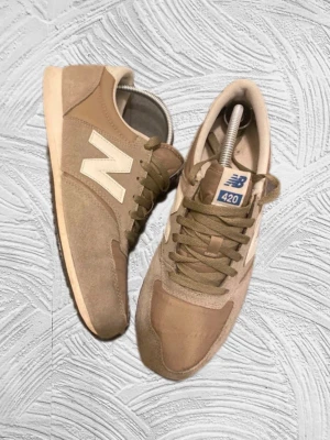 Beige New Balance 420 sneakers - Snygga beige New Balance 420 sneakers med klassisk vit N-logga på sidan. Skorna har en låg silhuett, ovandel i mocka och textil samt beige snörning. Perfekta för en avslappnad och trendig stil.