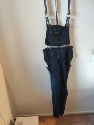 Mörkblå denim hängseljeans stl 42 - Unika mörkblå hängseljeans med bred frontficka, dekorativa knappar på sidorna och justerbara randiga hängslen. Jeansen har dragkedja i midjan och flera fickor. Perfekt för dig som vill sticka ut med en cool och retro look. Du är varmt välkommen att kika på mina andra annonser. 