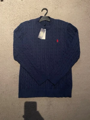 Mörkblå kabelstickad tröja Polo Ralph Lauren - Mörkblå kabelstickad tröja från Polo Ralph Lauren med rund halsringning och röd broderad logga på bröstet. Tröjan har långa ärmar och ribbade muddar vid ärmslut och nederkant. Perfekt för en stilren och klassisk look.